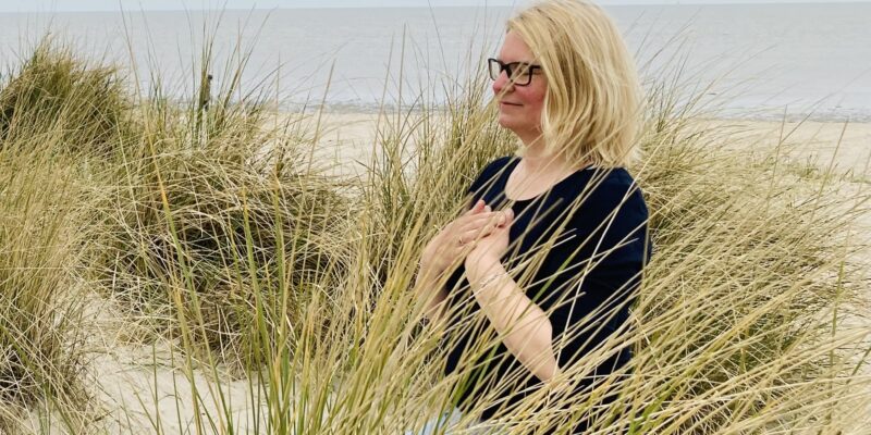 Frau kniet in den Dünen mit Händen auf dem Brustkorb, Meer im Hintergrund