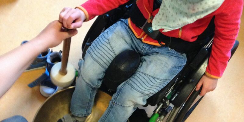 Kind im Rollstuhl von oben fotografiert, Füße in Klangschale, Schlegel gemeinsam gehalten