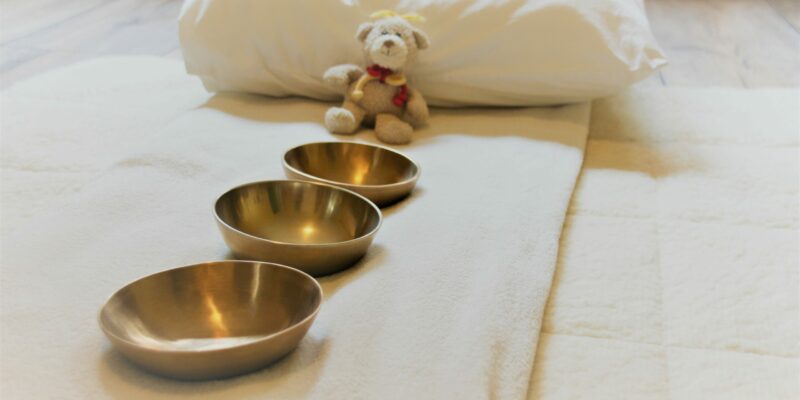 Klangraum für Kinder und Jugendliche Drei Klangschalen stehen vor einem kleinen Teddybären auf einer weichen Unterlage mit Kissen.