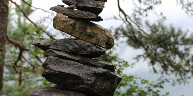 Steinstapel aus groben Steinen, symbolische Meilensteine, im Hintergrund eine luftige Tanne und weißer Himmel.