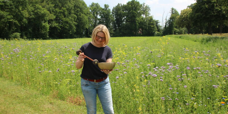 Klangmassage in der Natur Frau hält eine Klangschale auf einer Sommerwiese und schlägt sie sanft mit einem Schlegel an