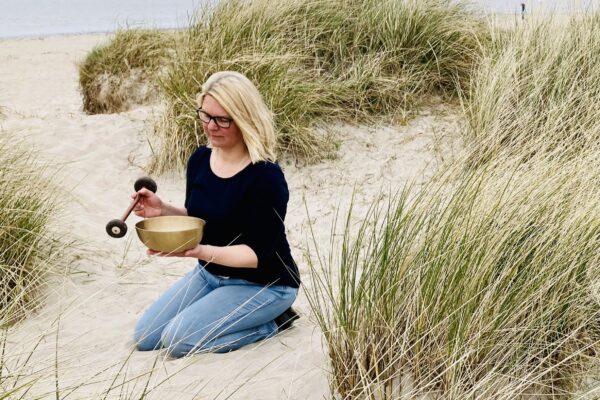 Klangschale zwischen Dünen – Klangmassage in Verl Frau kniet zwischen Dünen in Verl, hält eine Klangschale und einen Schlägel in den Händen, blickt in die Klangschale, trägt dunkles Oberteil und Jeans, blonde offene Haare, Brille – Wohlfuehlzeit Verl.