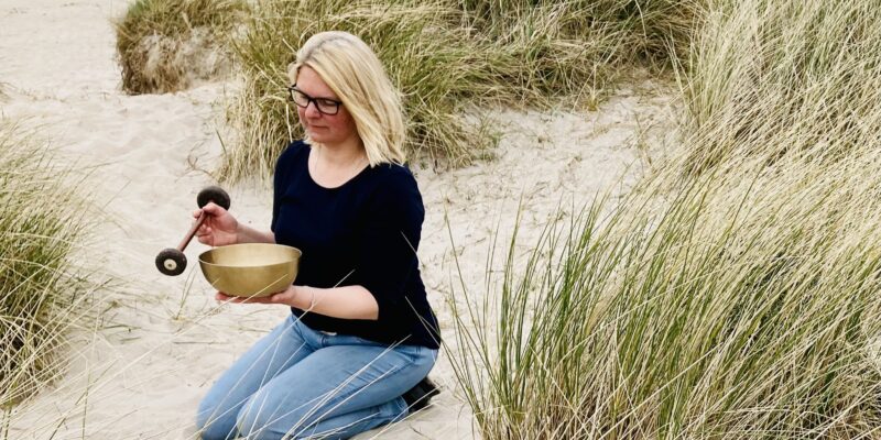 Frau kniet zwischen Dünen in Verl, hält eine Klangschale und einen Schlägel in den Händen, blickt in die Klangschale, trägt dunkles Oberteil und Jeans, blonde offene Haare, Brille – Wohlfuehlzeit Verl.