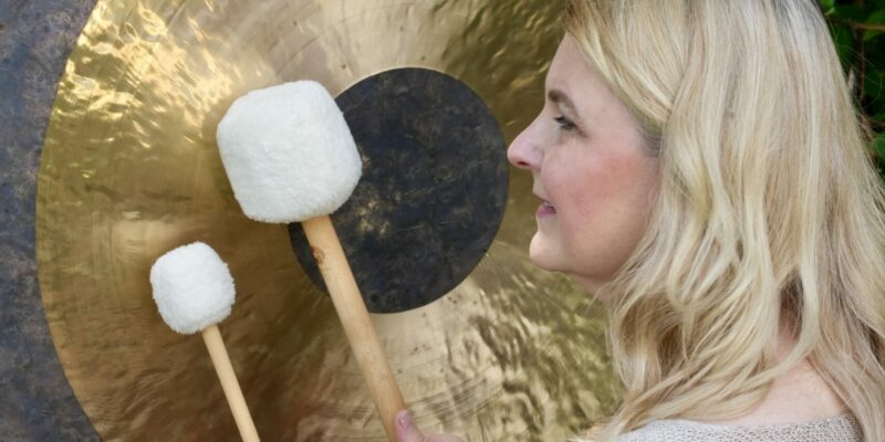 Frau kniet vor einem schwarz-goldenen Tam-Tam-Gong in der Natur, hält zwei unterschiedlich große Schlägel mit weißem Filzkopf, blickt seitlich zum Gong, trägt beigen Pullover und hat blondes, halblanges offenes Haar. Wohlfuehlzeit Verl.