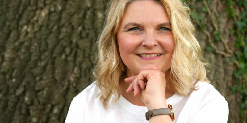 Frau vor Baum, Oberkörper sichtbar, lächelnd in die Kamera – Coaching, Achtsamkeit und orthopädische Yogatherapie, Yoga und Begleitung in Verl – Porträt von Ulrike Braak“