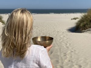 Frau am Strand mit Klangschale in der Hand blickt Richtung Meer und Horizont