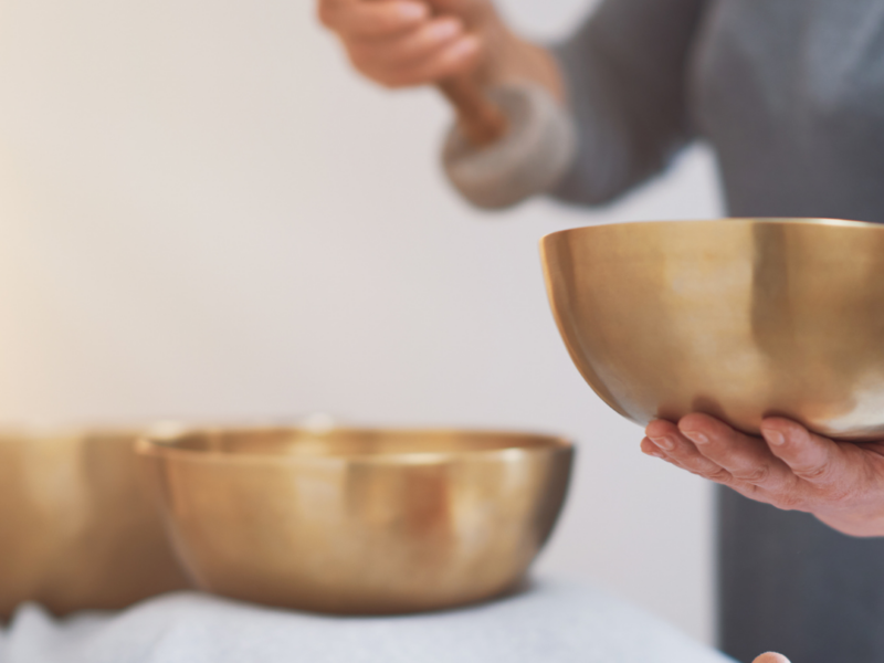 Frau liegt auf einer Liege, auf Brustkorb und Bauch stehen zwei goldene Klangschalen, daneben steht eine weitere Frau und hält eine Klangschale sowie einen Holzschlägel mit Filzkopf.