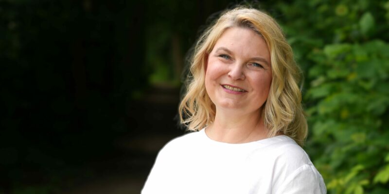 Porträt von Ulrike Braak am Waldrand, lächelnd in die Kamera, helles weißes Oberteil, schemenhafter Weg im dunklen Hintergrund, grünes Umfeld.