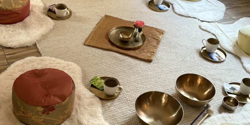 Yogaraum mit Sitzplätzen auf Yogakissen, Klangschalen und gestalteter Mitte, jeweils Tablett mit Tasse und Kärtchen