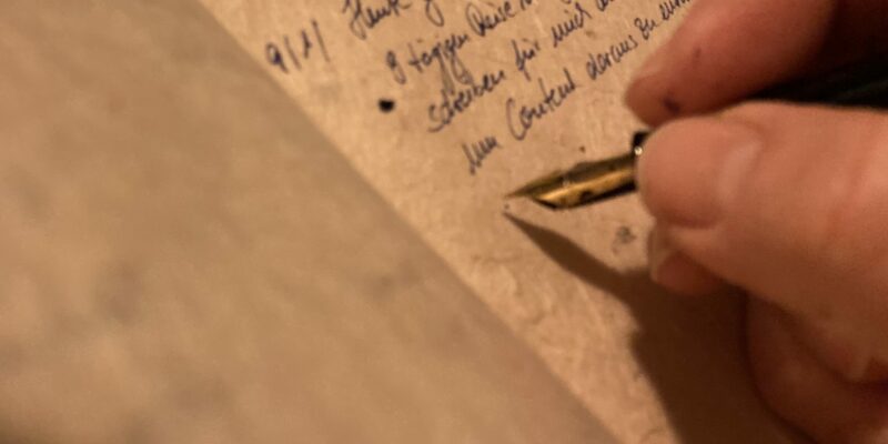 Aufgeschlagenes Buch mit handgeschriebenen Zeilen auf Naturpapier, Füller in Hand, nur Daumen und Zeigefinger sichtbar