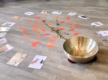 Symbolische Klangschale – Fuchs-Geschichte über Trauer Große goldene Klangschale auf Holzfußboden, umgeben von Zweig und orangefarbenen Papierblättern wie Baumwurzel, Bilder aus der Geschichte vom Fuchs und seinen Waldtieren über Sterben und Trauer – Wohlfuehlzeit Verl.