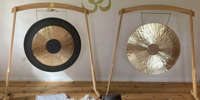 Das Foto zeigt eine goldene Klangschale am Strand in Verl, gehalten von einer nur leicht sichtbaren Hand rechts unten im Bild. Links ist Dünengras zu sehen, im Hintergrund erstrecken sich Strand und Meer unter einem hellblau-grauen Himmel. Das Bild vermittelt Ruhe, Achtsamkeit und die besondere Atmosphäre der Klangpraxis in offenen Klangräumen und am Meer, wie sie bei Wohlfuehlzeit-Verl.de angeboten wird.
