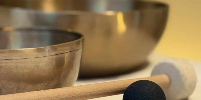 Zwei Klangschalen auf weißer Unterlage, davor zwei Schlegel mit Holzgriff (schwarze Kugel und weißer Filzring), im Hintergrund schemenhaft ein Tam-Tam-Gong.