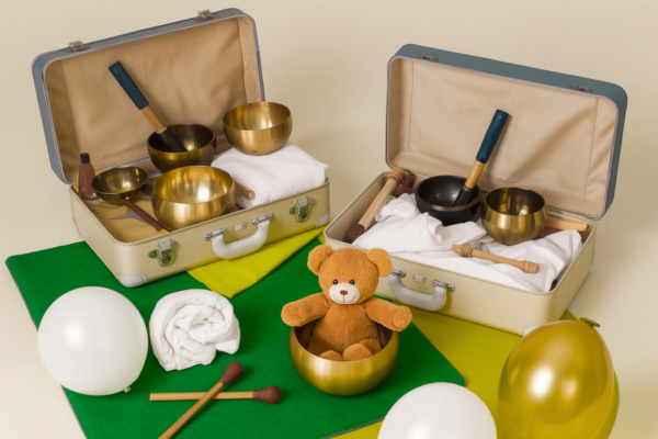 KinderKlangInsel – Klangmaterialien und mobile Klangwelt Zwei geöffnete helle Koffer mit Klangschalen, weißen Decken und Schlägeln. Darunter liegen eine grüne und eine gelbe Matte. Weiße und gelbe Luftballons sind im Bild verteilt. Vor den Koffern steht eine goldene Klangschale mit einem kleinen Teddybären darin. Davor liegen zwei Schlägel.