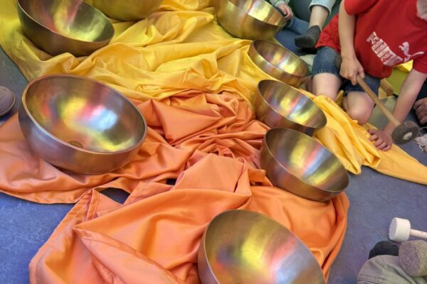 KinderKlangInsel – Klang erleben mit Schwingung, Farbe und Wahrnehmung Klangschalen stehen auf orangenen Tüchern, die in Regenbogenfarben schimmern. Um die Klangschalen sitzen Kinder auf dem Boden, sichtbar sind nur Beine und Rumpf sowie Hände mit Klangschlägeln. Die Szene zeigt ein ruhiges Klangangebot im Raum auf Bodenniveau.
