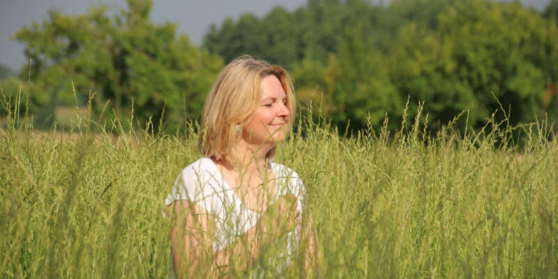 Frau mit blonden Haaren sitzt in einer hohen Wiese vor einem Wald und genießt mit geschlossenen Augen die Sonne.