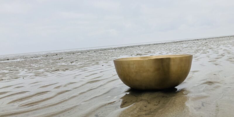 Klangschale am Meer – Trauer braucht Wärme Klangschale auf dem Sandstrand bei Ebbe, mit flachen Wasserspuren im Sand und bewölktem Himmel.